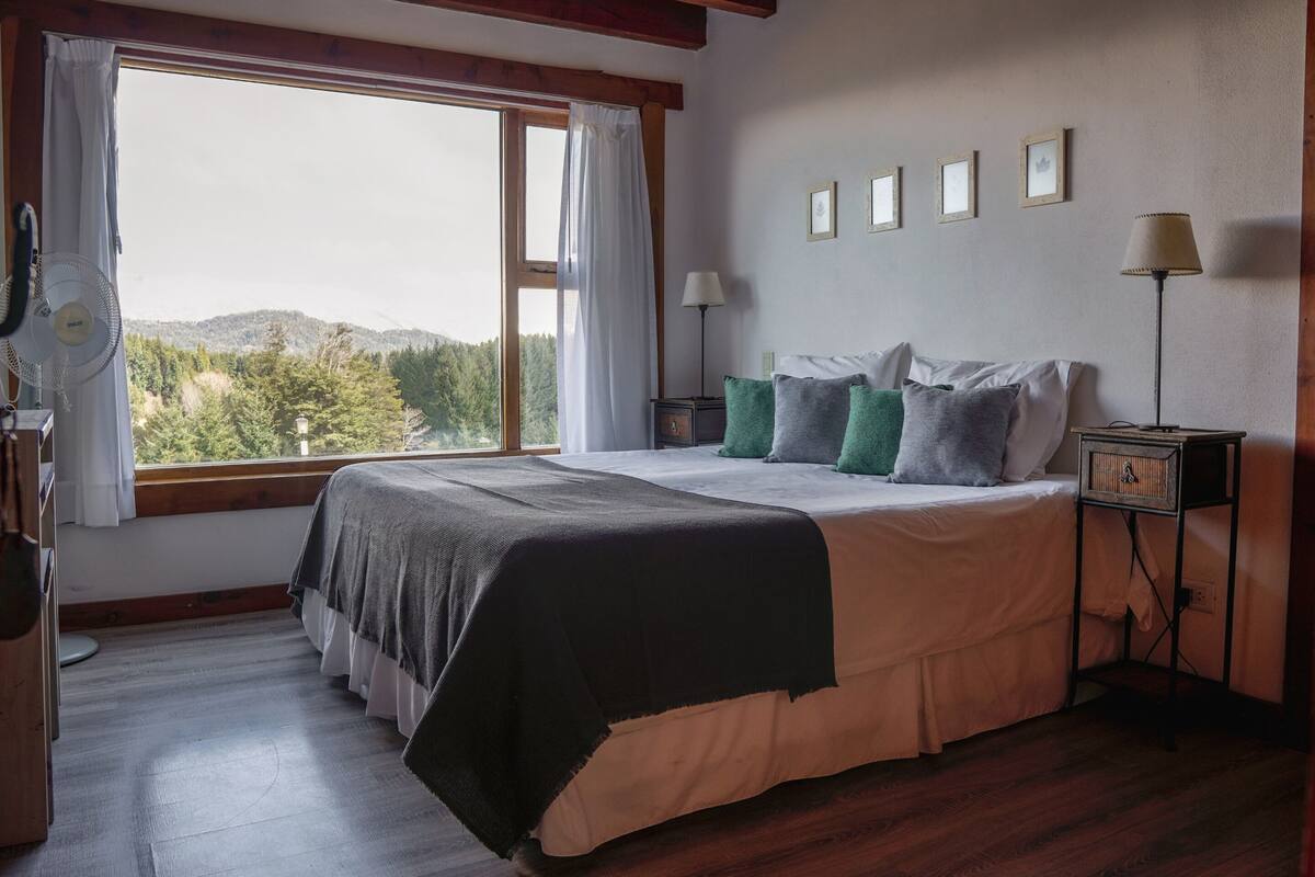 Une chambre avec un grand lit, une fenêtre avec des rideaux, une table de chevet avec une lampe, et une vue sur l’extérieur.