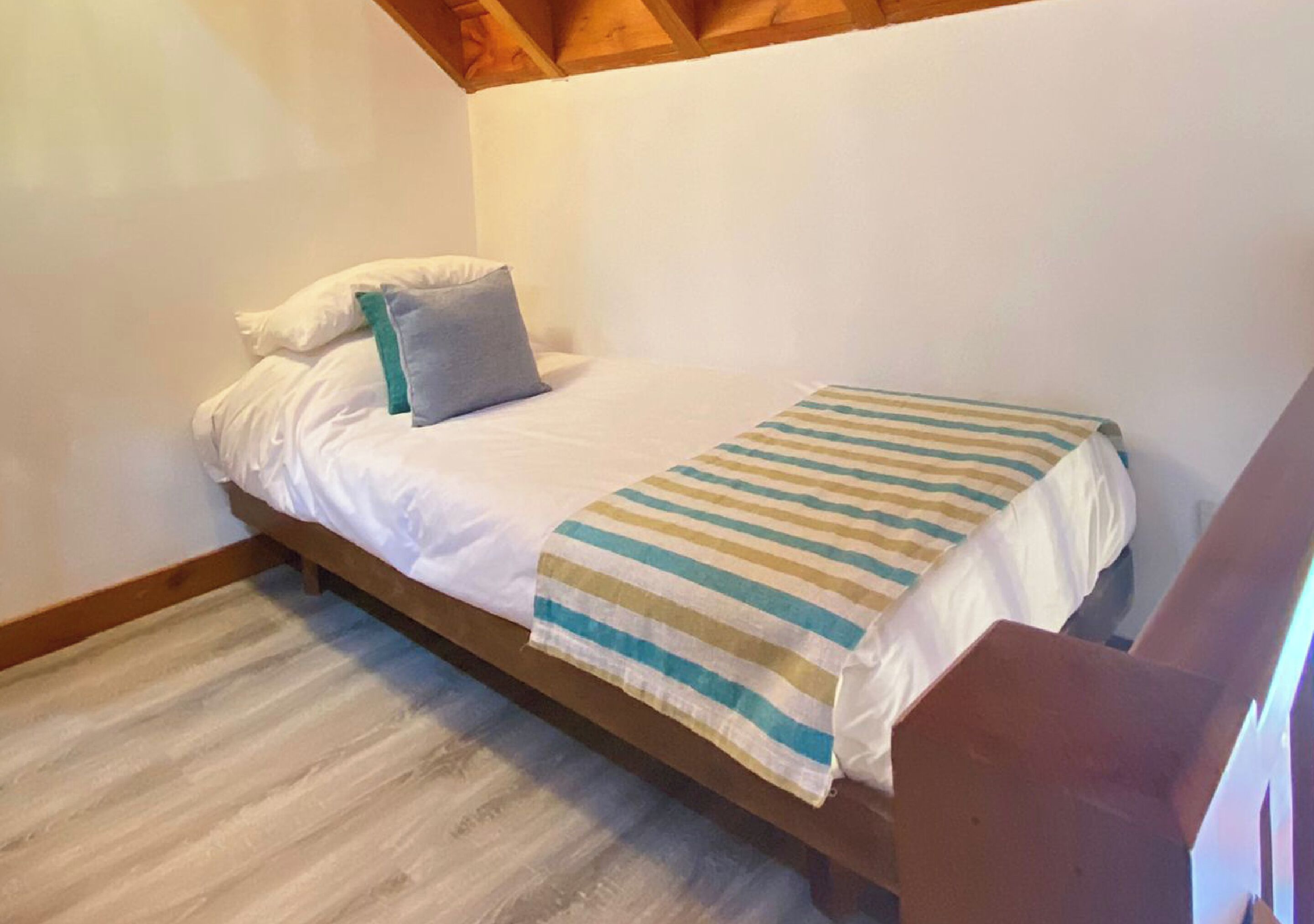 Uma cama de solteiro com manta listrada, almofadas brancas e cabeceira de madeira, em quarto com piso e paredes brancas.