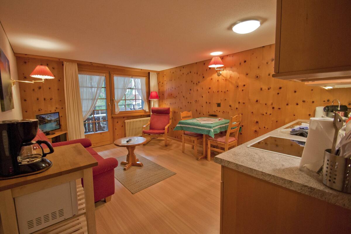Una habitación acogedora con piso de madera, una mesa de comedor con sillas, una pequeña mesa de centro, un sillón rojo y un televisor sobre un mueble de madera.