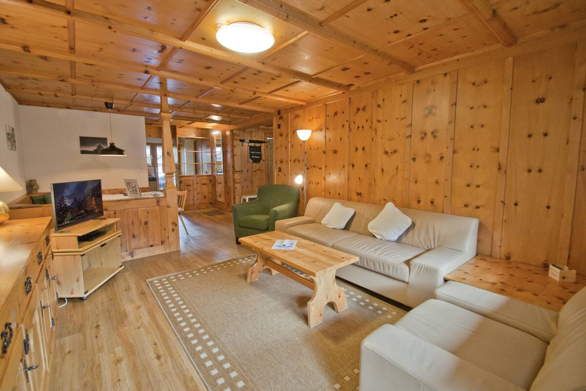 Interior de una cabaña de madera con un sofá, una silla verde, una mesa de madera y un televisor sobre un mueble de madera.