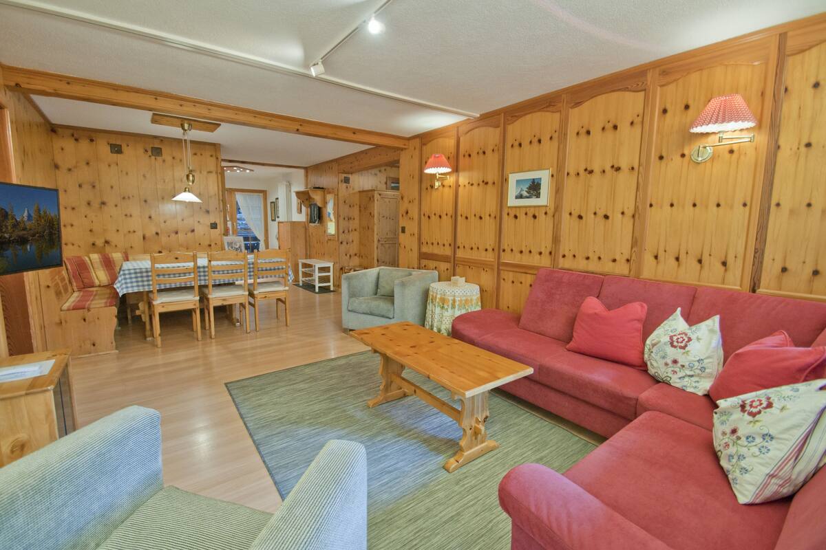 Un salón acogedor con paredes de madera, un sofá rojo, una mesa de centro de madera y una zona de comedor con mesa y sillas.