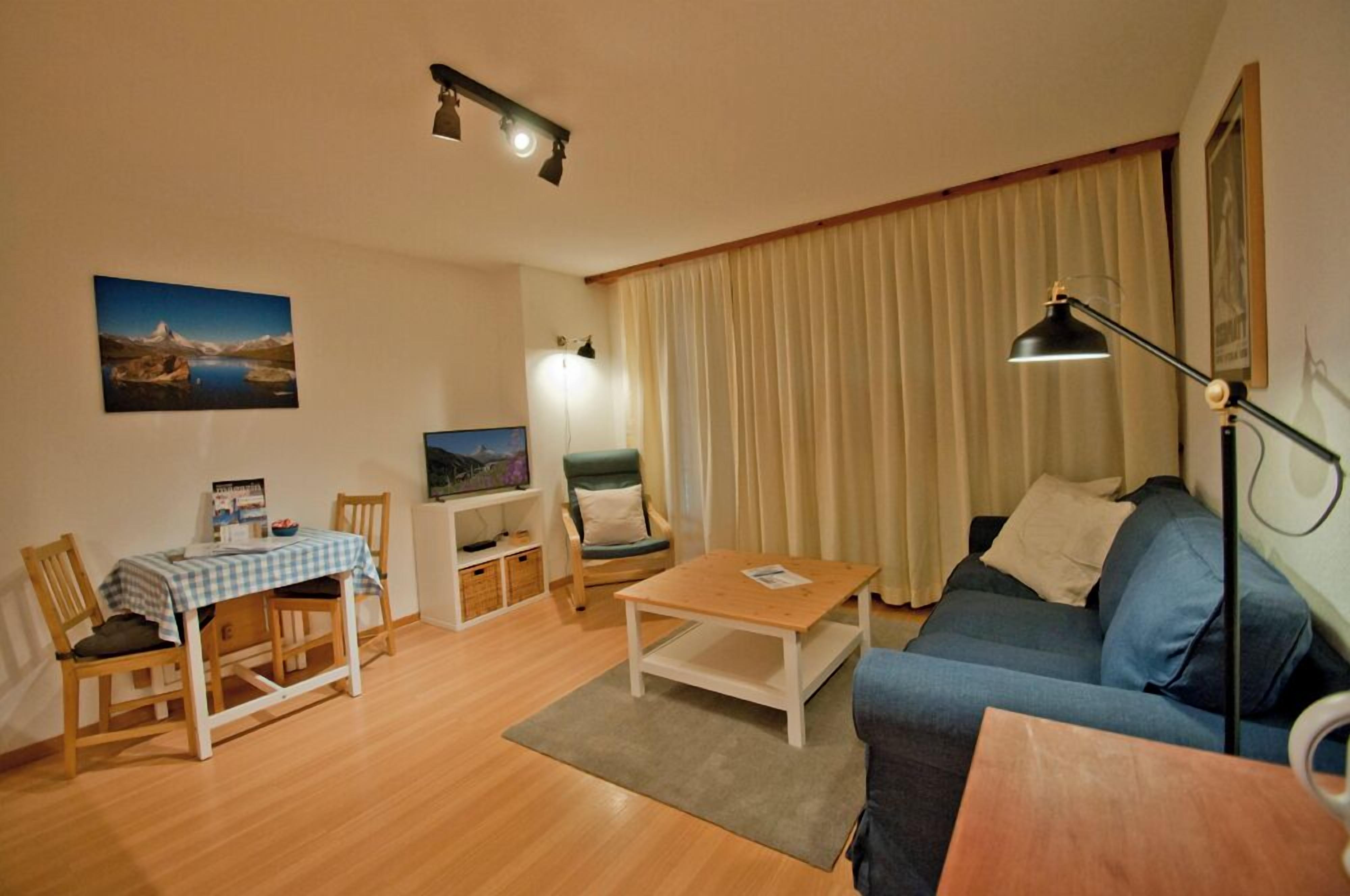 Een gezellige woonkamer met een blauwe bank, een houten salontafel, een eettafel met stoelen, een televisietoestel en een lamp.