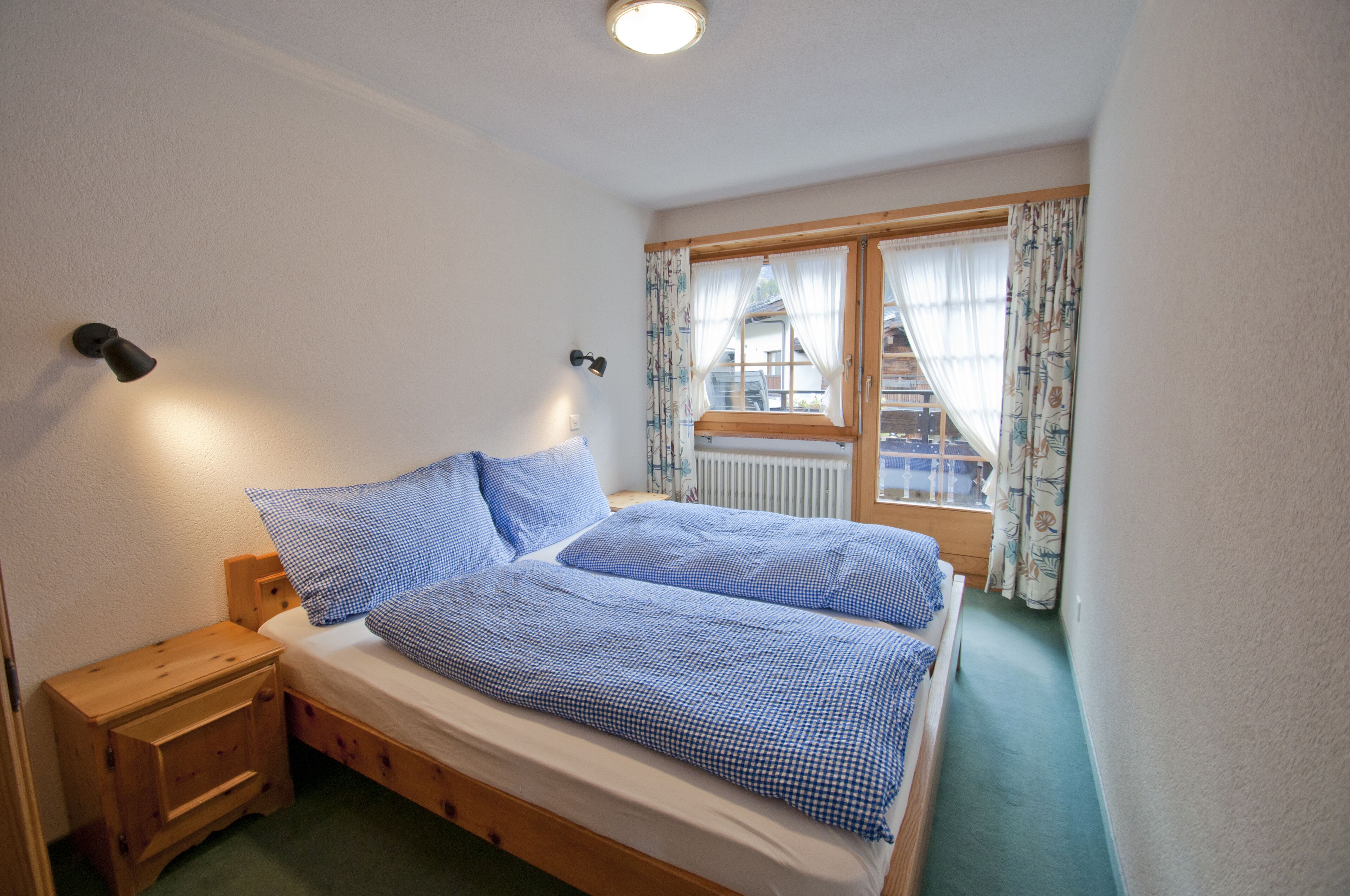 Une chambre à coucher avec un lit en bois, une table de chevet, une fenêtre avec des rideaux et une vue sur un bâtiment à l’extérieur.