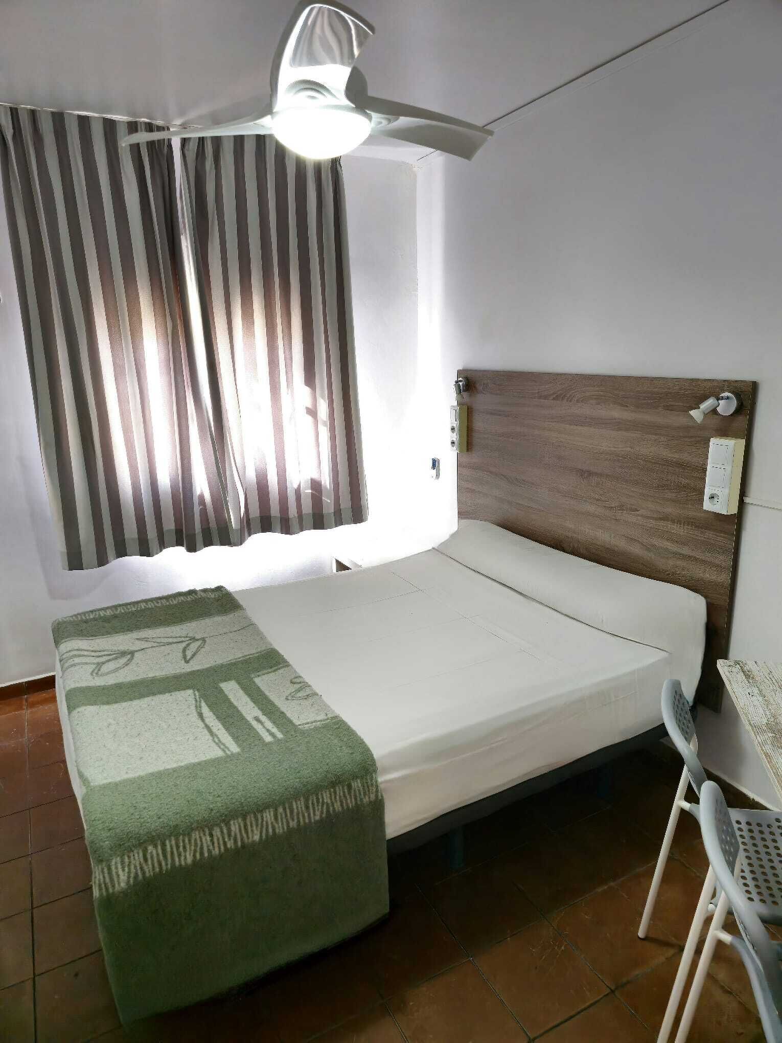 Une chambre avec un lit simple, une tête de lit en bois, un ventilateur de plafond, une fenêtre avec des rideaux à rayures et une petite table avec une chaise.