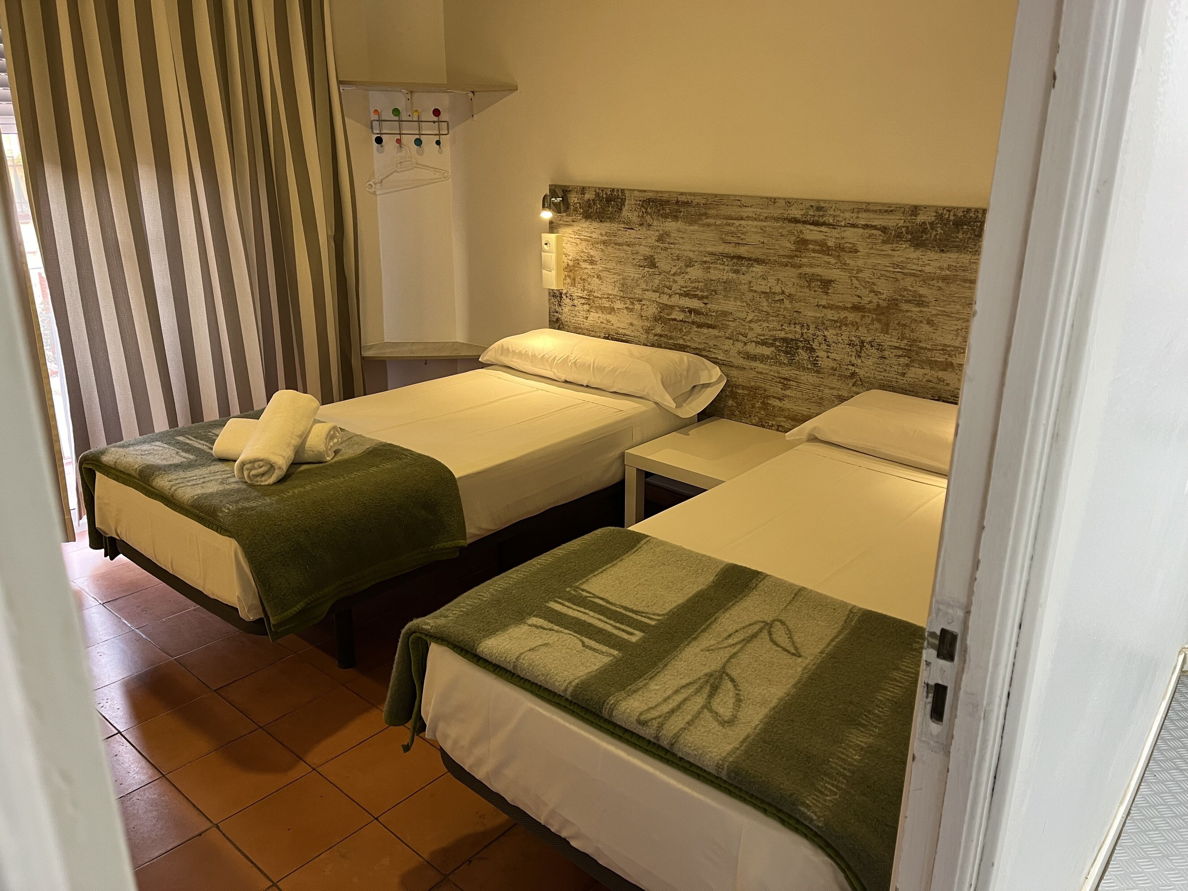 Camera d'albergo con due letti singoli, ognuno con un asciugamano verde e lenzuola bianche. Tra i letti si trova un comodino piccolo, e un armadio bianco con uno specchio sopra.