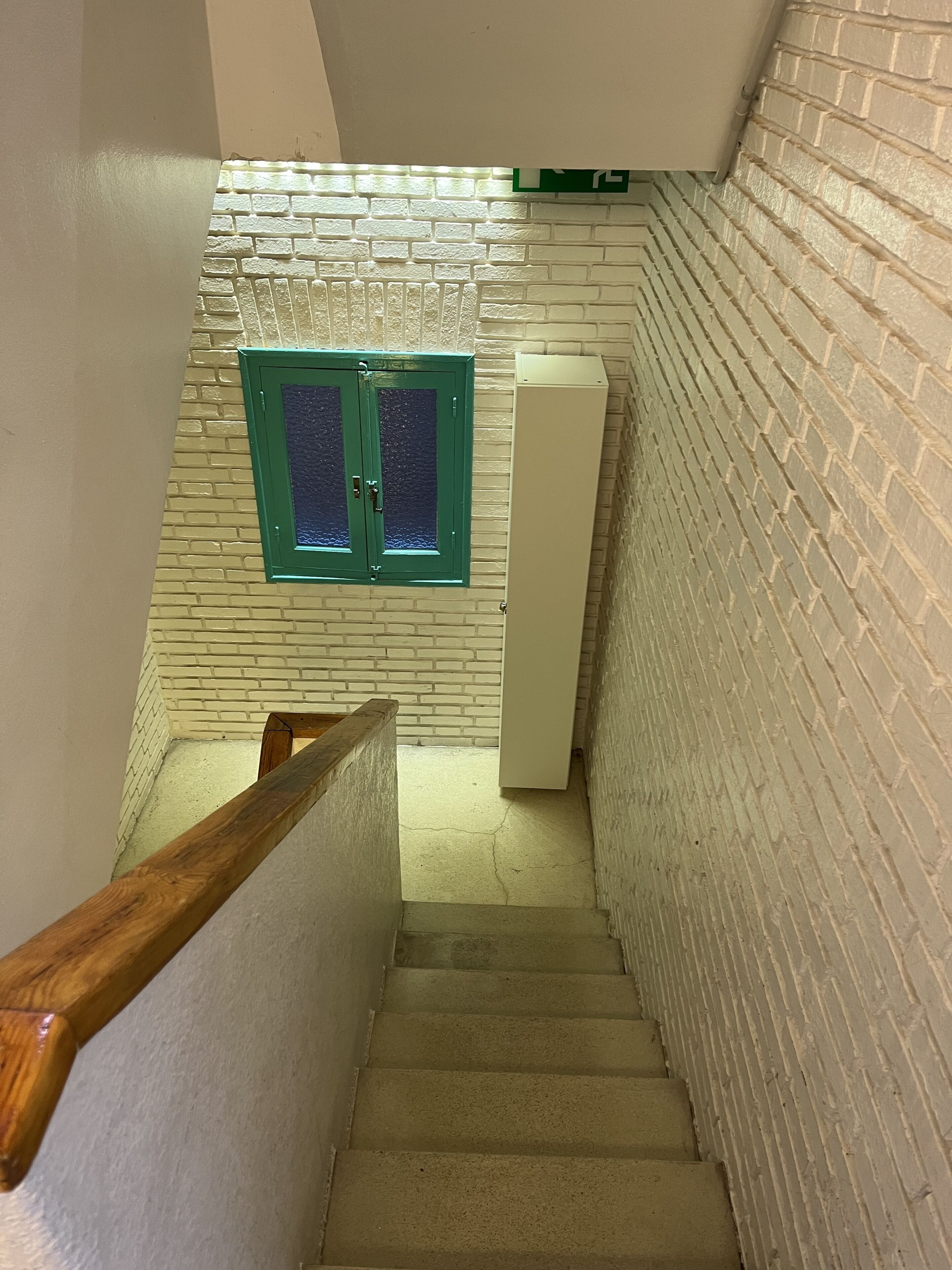 Una escalera con barandilla de madera, un ventanal con marco verde y una pared blanca con diseño de ladrillo.