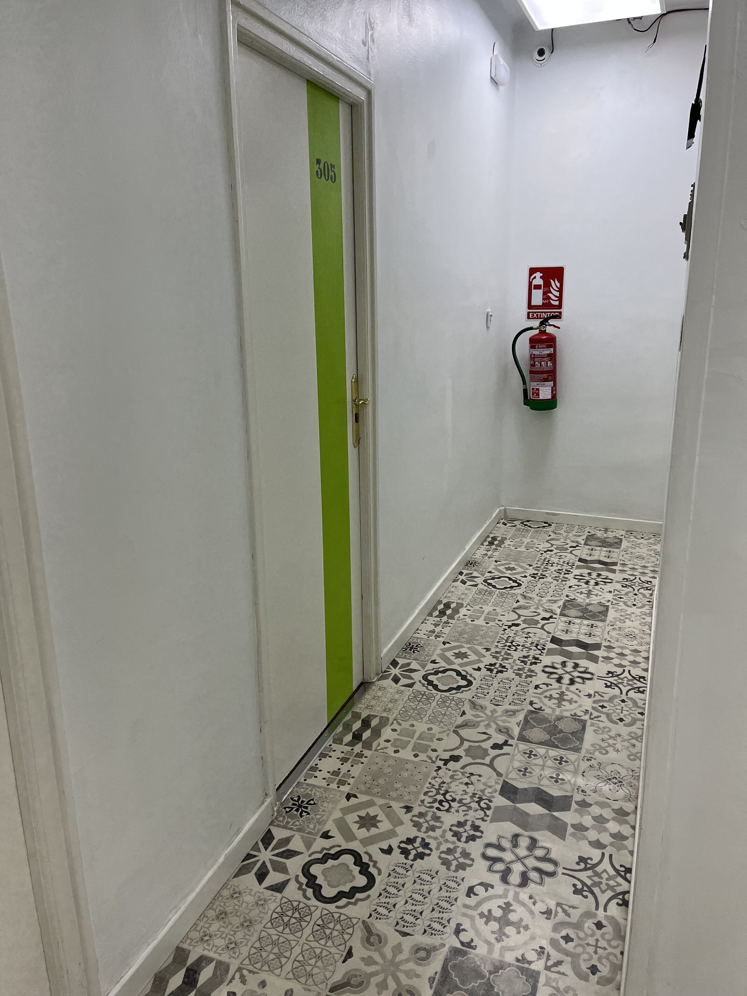 Ein Flur mit gemusterten Bodenfliesen, einer grünen Tür mit der Beschriftung '305' und einem an der Wand befestigten Feuerlöscher.