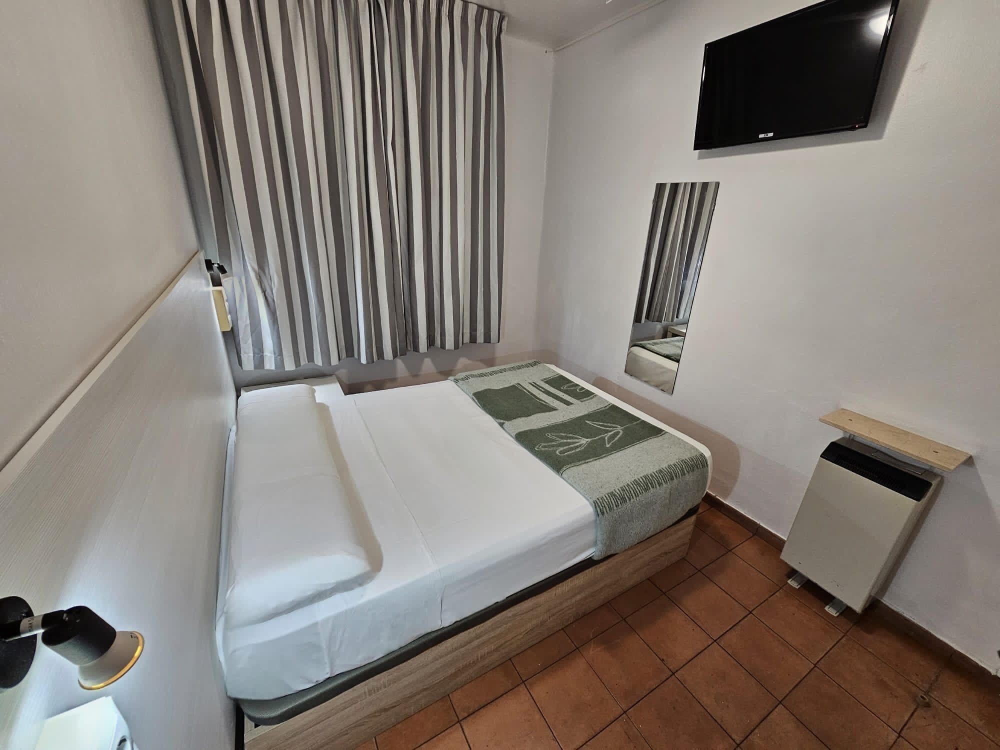 Una cama individual con colchón blanco y una manta verde y blanca, una mesita de noche pequeña, un televisor montado en la pared y una ventana con persianas verticales.