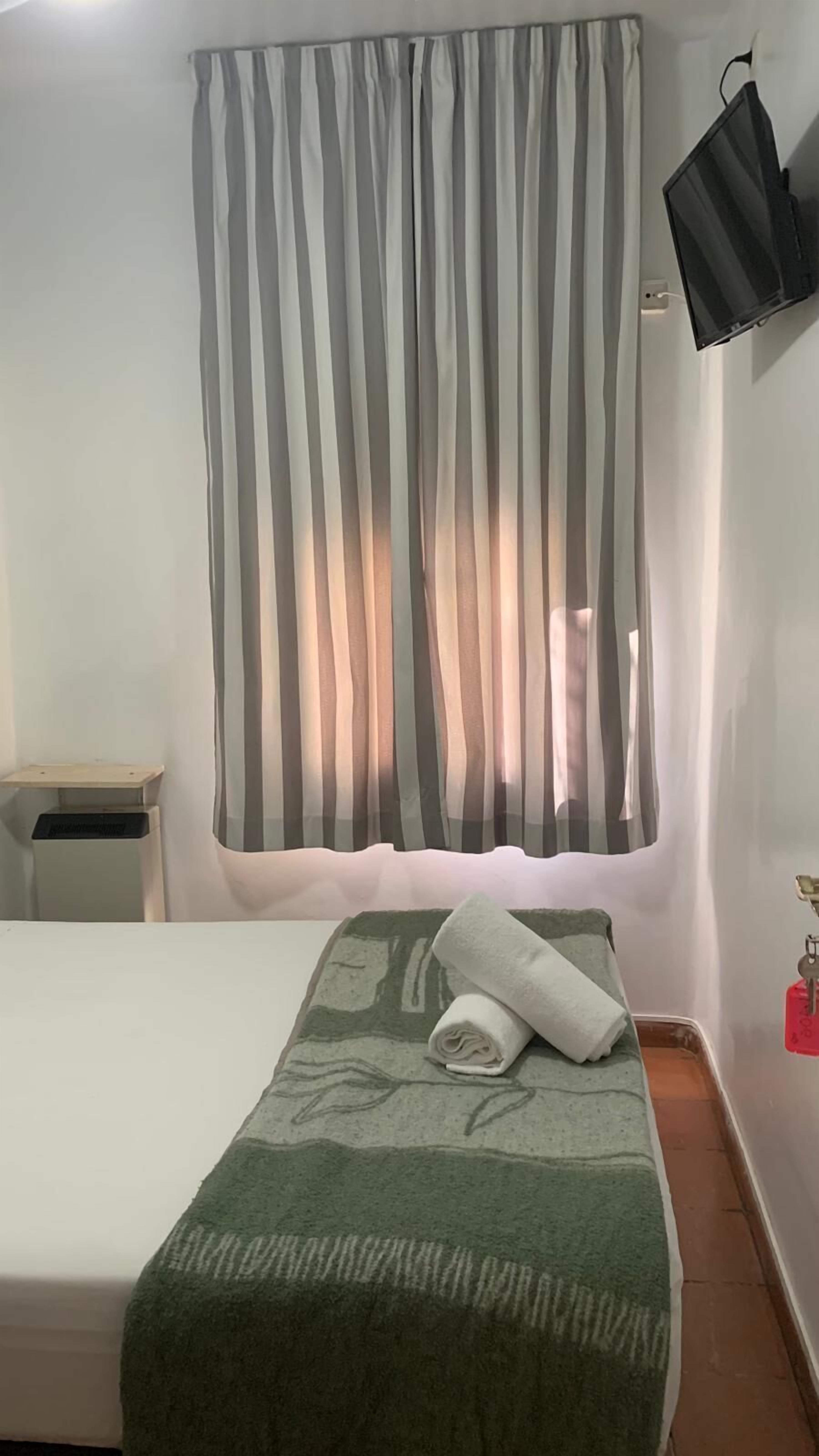 A room with a bed, a green blanket, a striped curtain, a small table, and a wall-mounted TV.