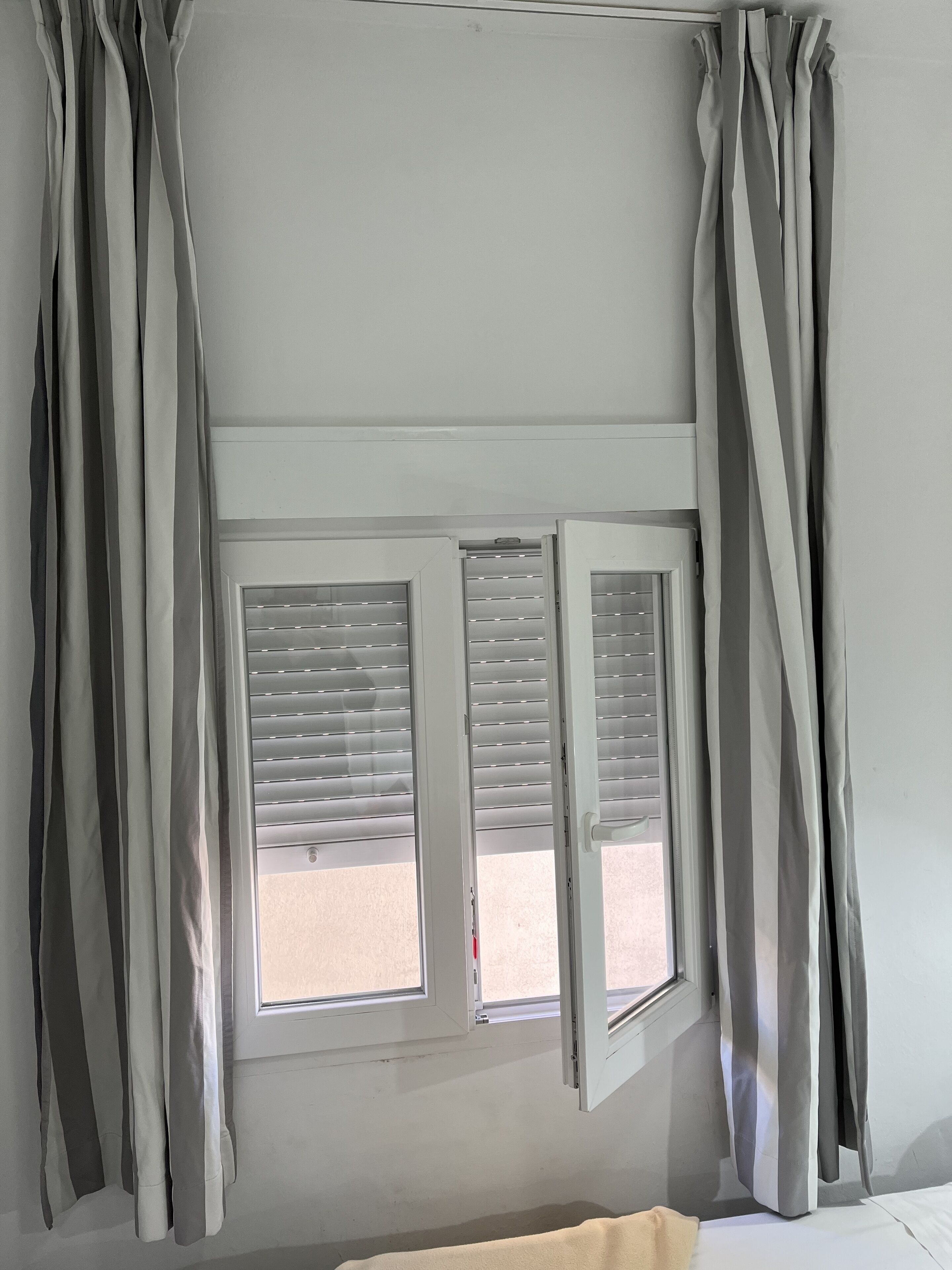 A room with a window featuring horizontal blinds, a bed with a white cover, and grey curtains.