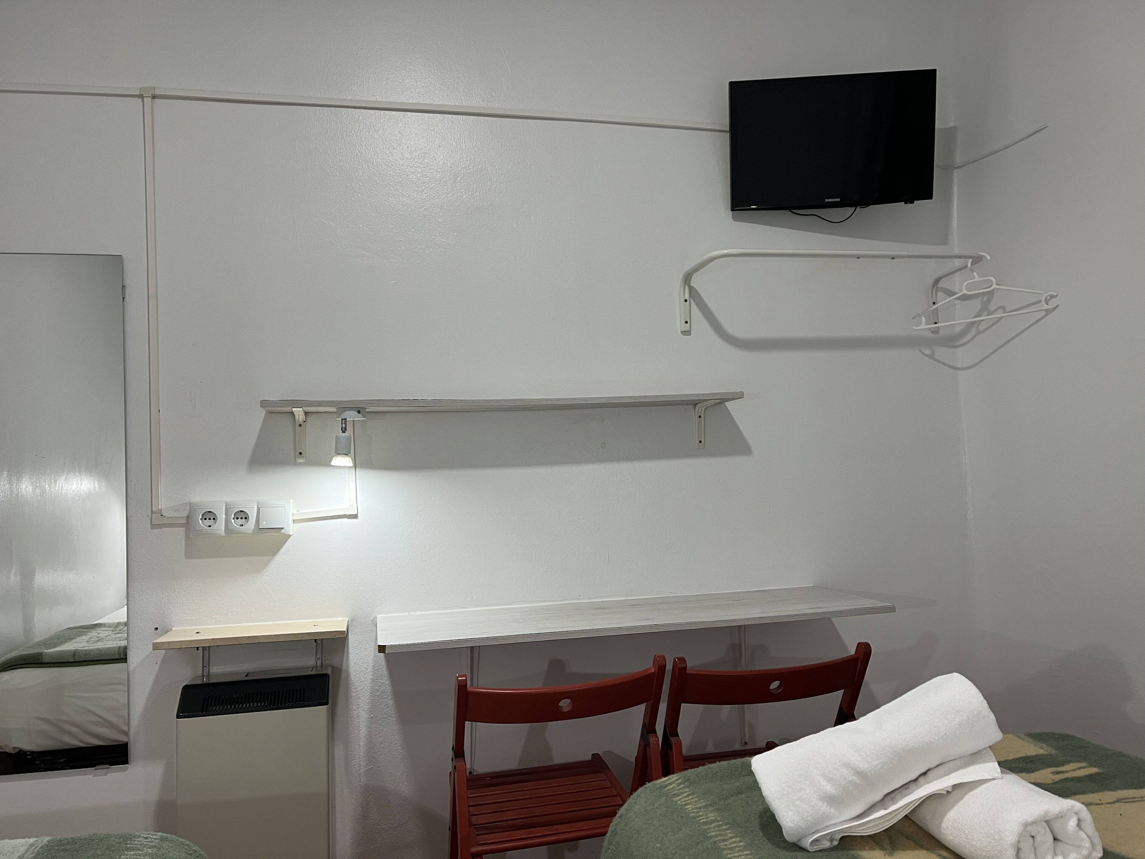 A small, simple room with a bed, a desk, a chair, a TV mounted on the wall, and a shelf.