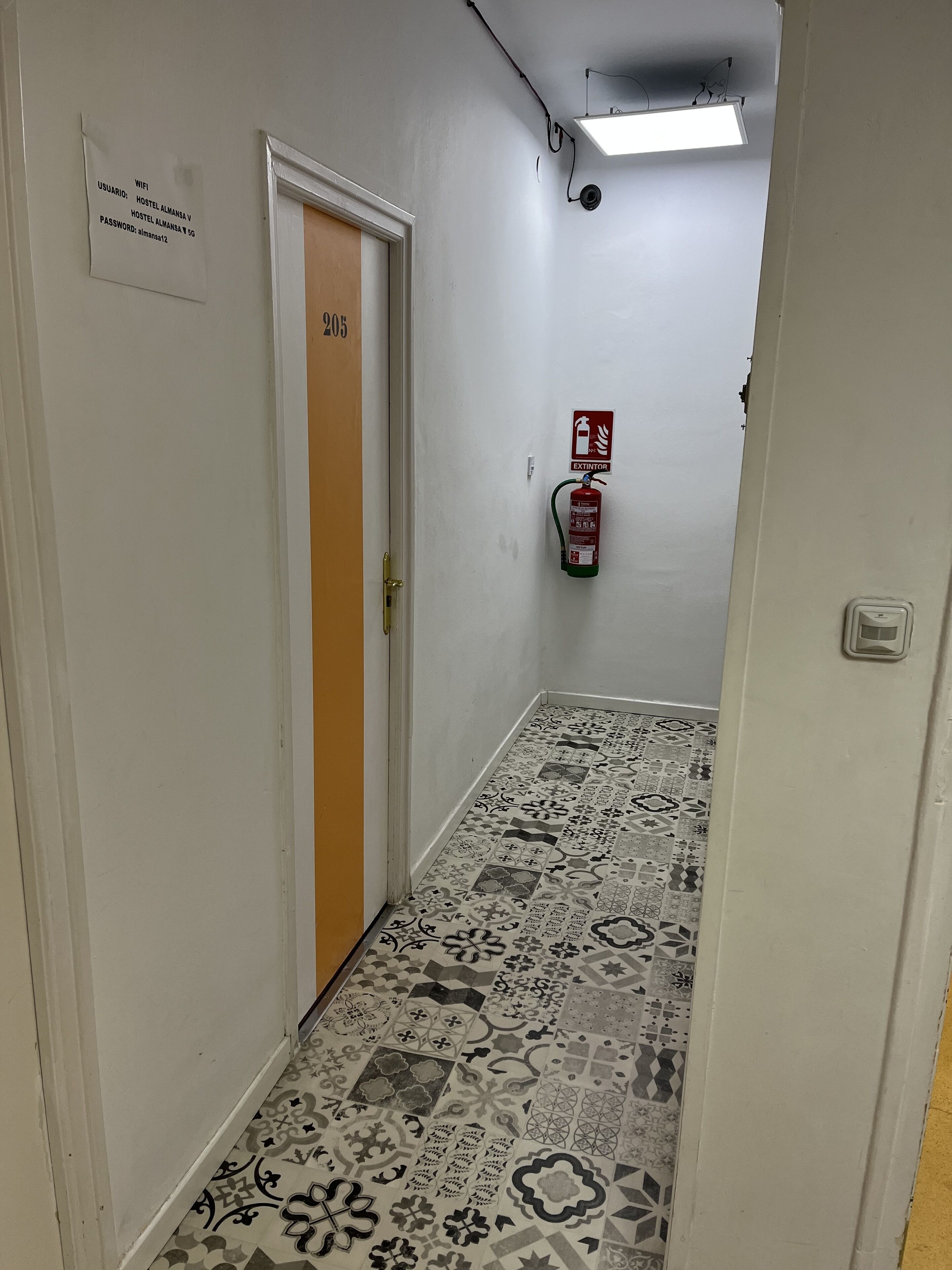 Een hal met vloertegels met een patroon, een deur met de aanduiding '205', een brandblusser en een bord met wifi-gegevens.