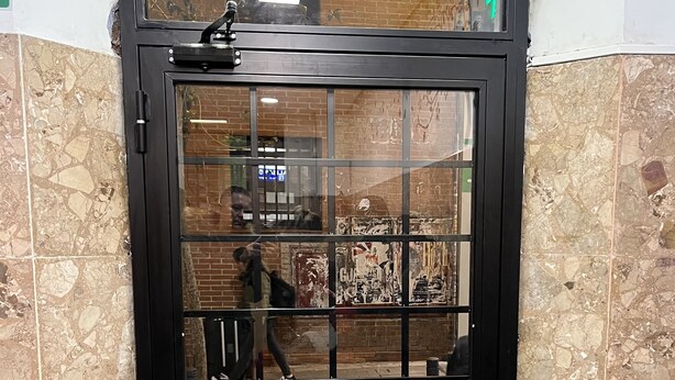 A black metal gate with a glass panel, set in a corner with a tiled floor and a brick wall. - Casa de huespedes Almansa (Madrid)