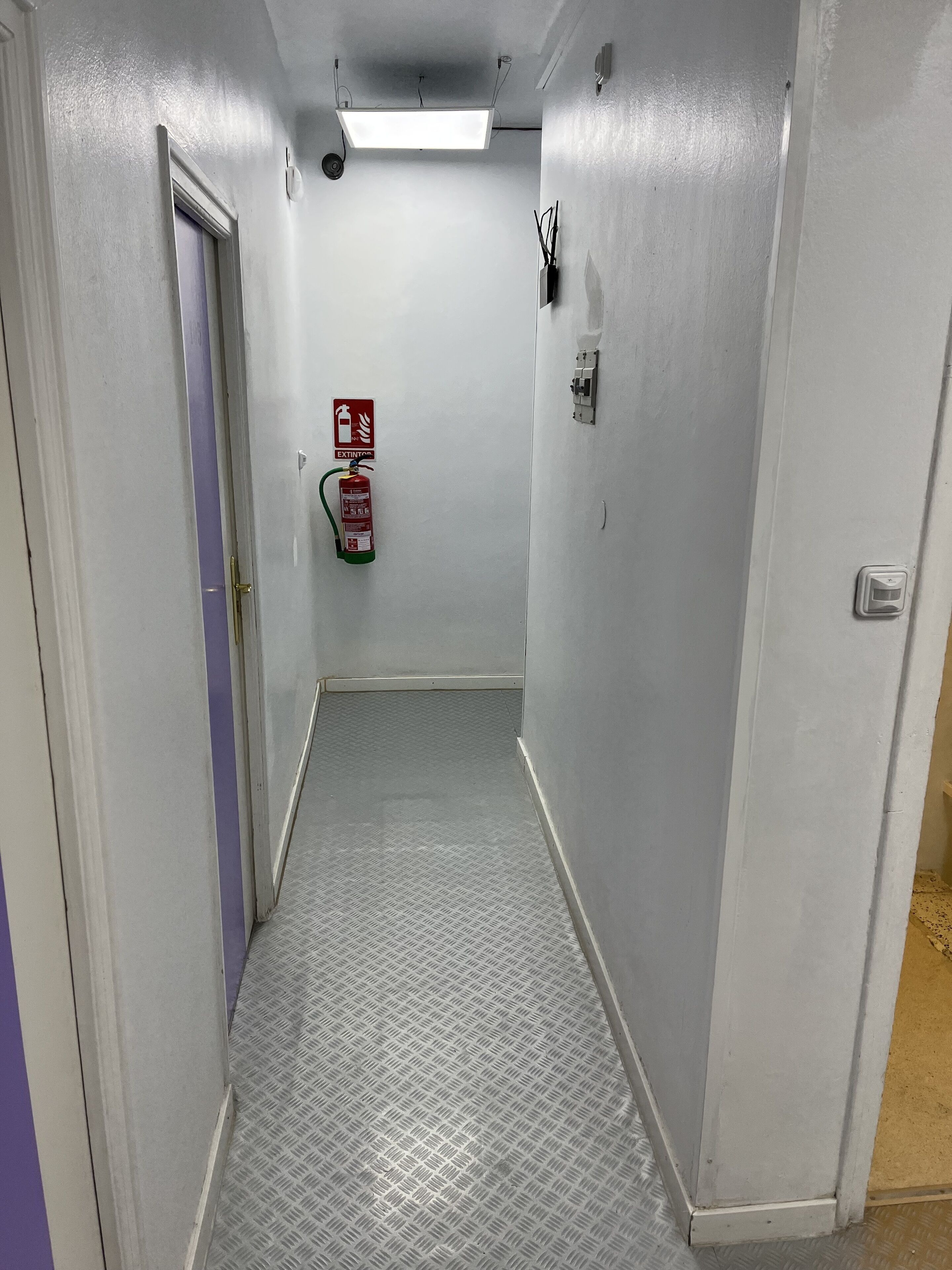 Un couloir étroit aux murs blancs, un extincteur fixé au mur et une porte ornée d’une bande violette.