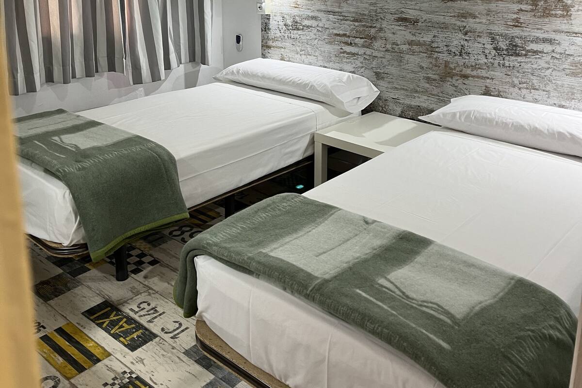A hotel room with two single beds, a headboard with a textured wall, a window with curtains, and a patterned floor.