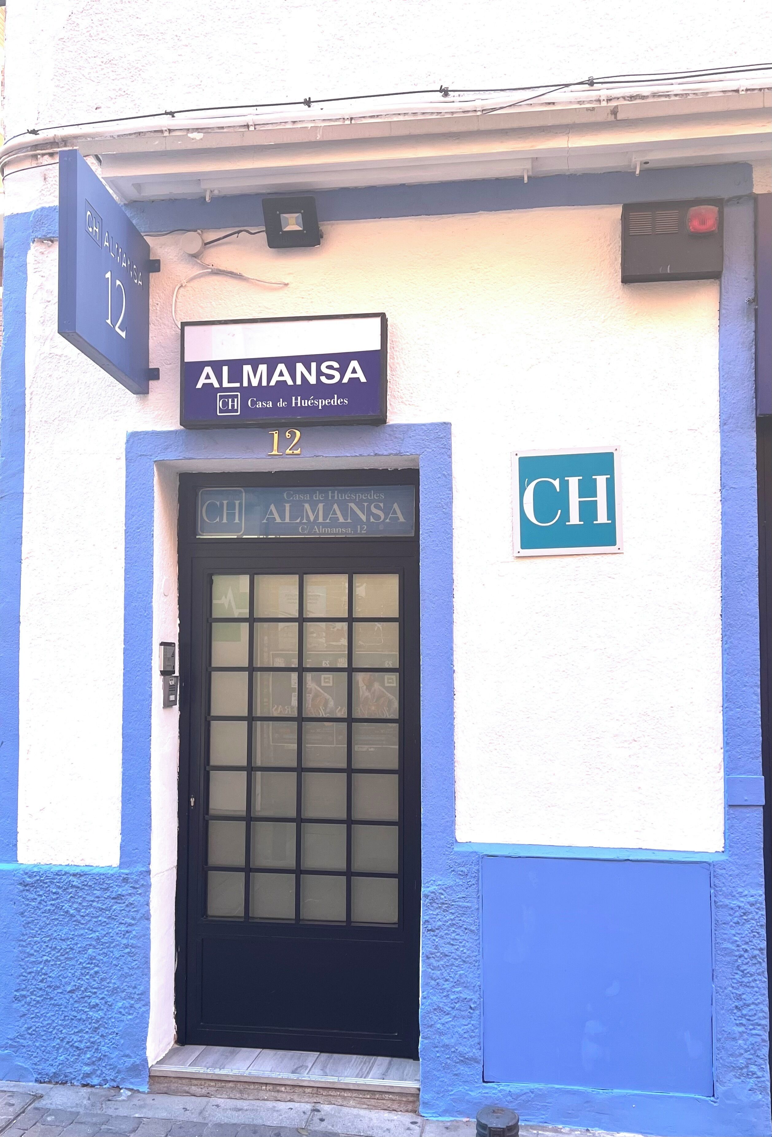 A building facade with a blue and white exterior, a black door with glass panels, and signs indicating 'ALMANSA' and 'CH'.
