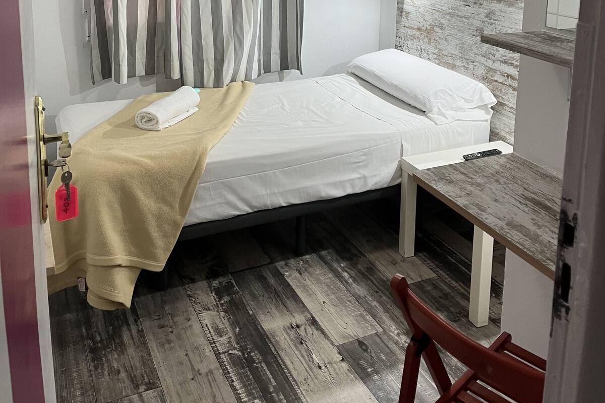 A room with a bed, a desk, a chair, a ceiling fan, and a curtain.