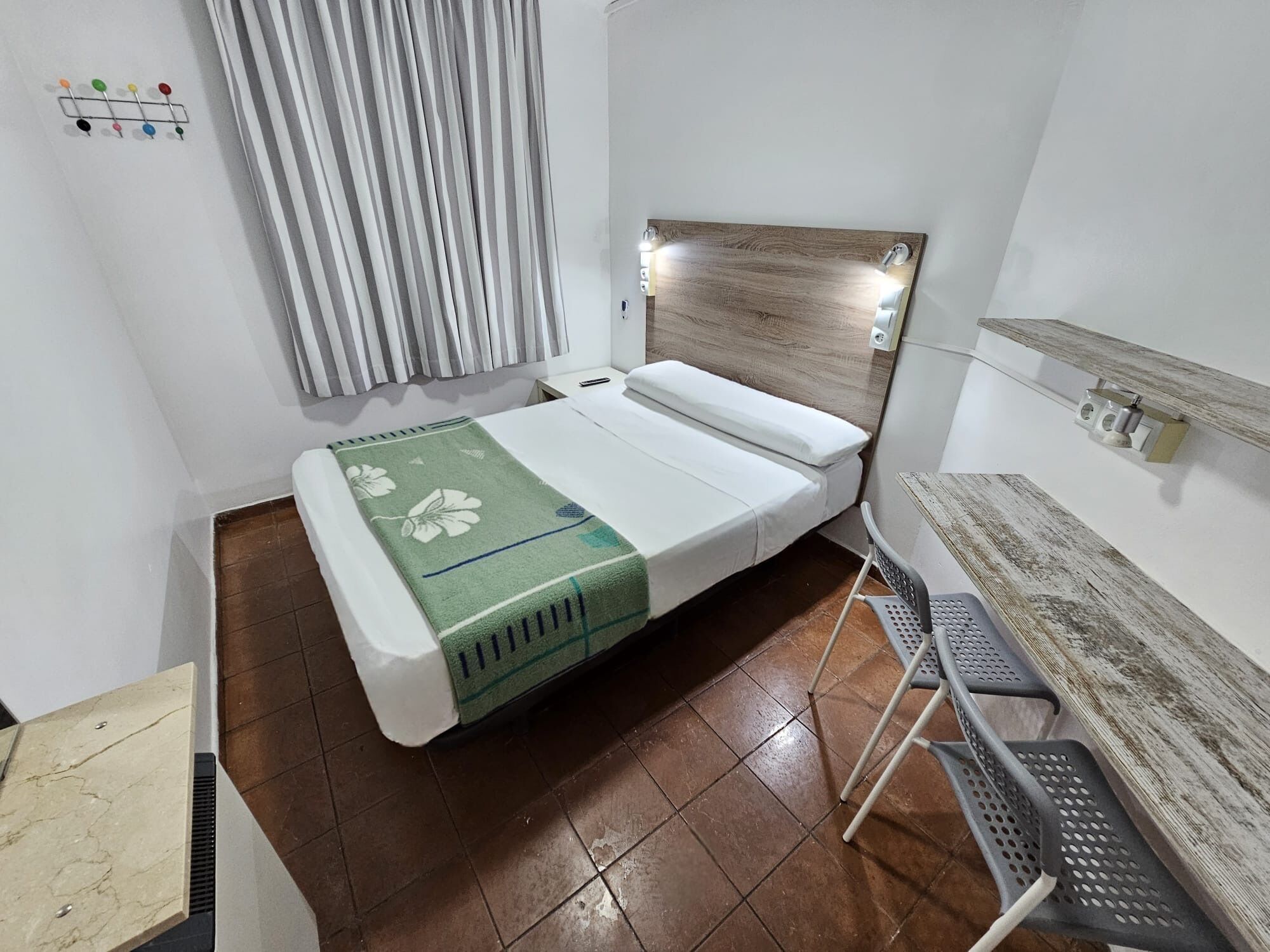 A small, neatly arranged room with a bed, a chair, a shelf with items, and a curtain.