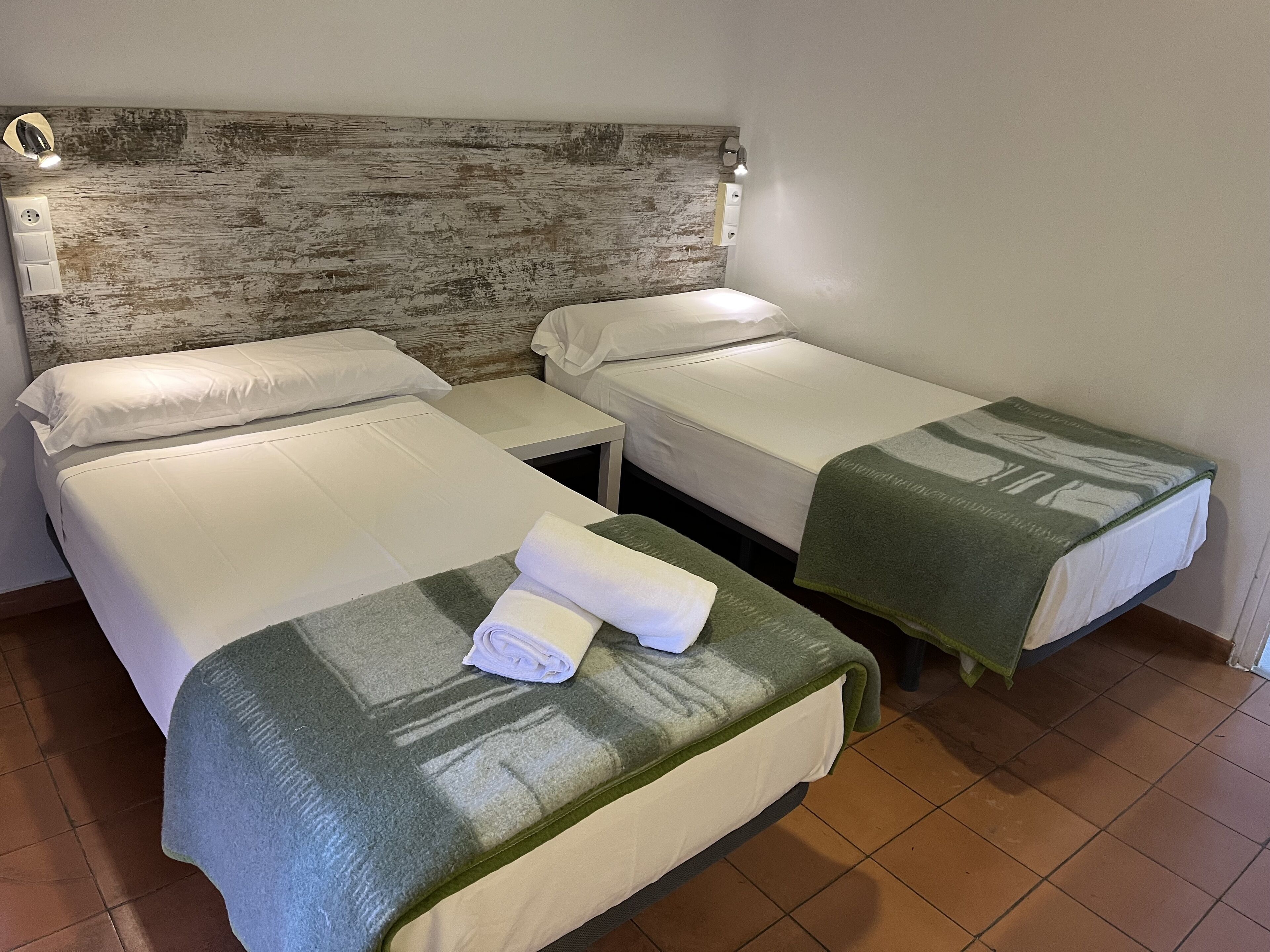 Habitación de hotel con dos camas individuales, una mesita de noche pequeña, una lámpara fijada a la pared y un cabecero de madera rústica.
