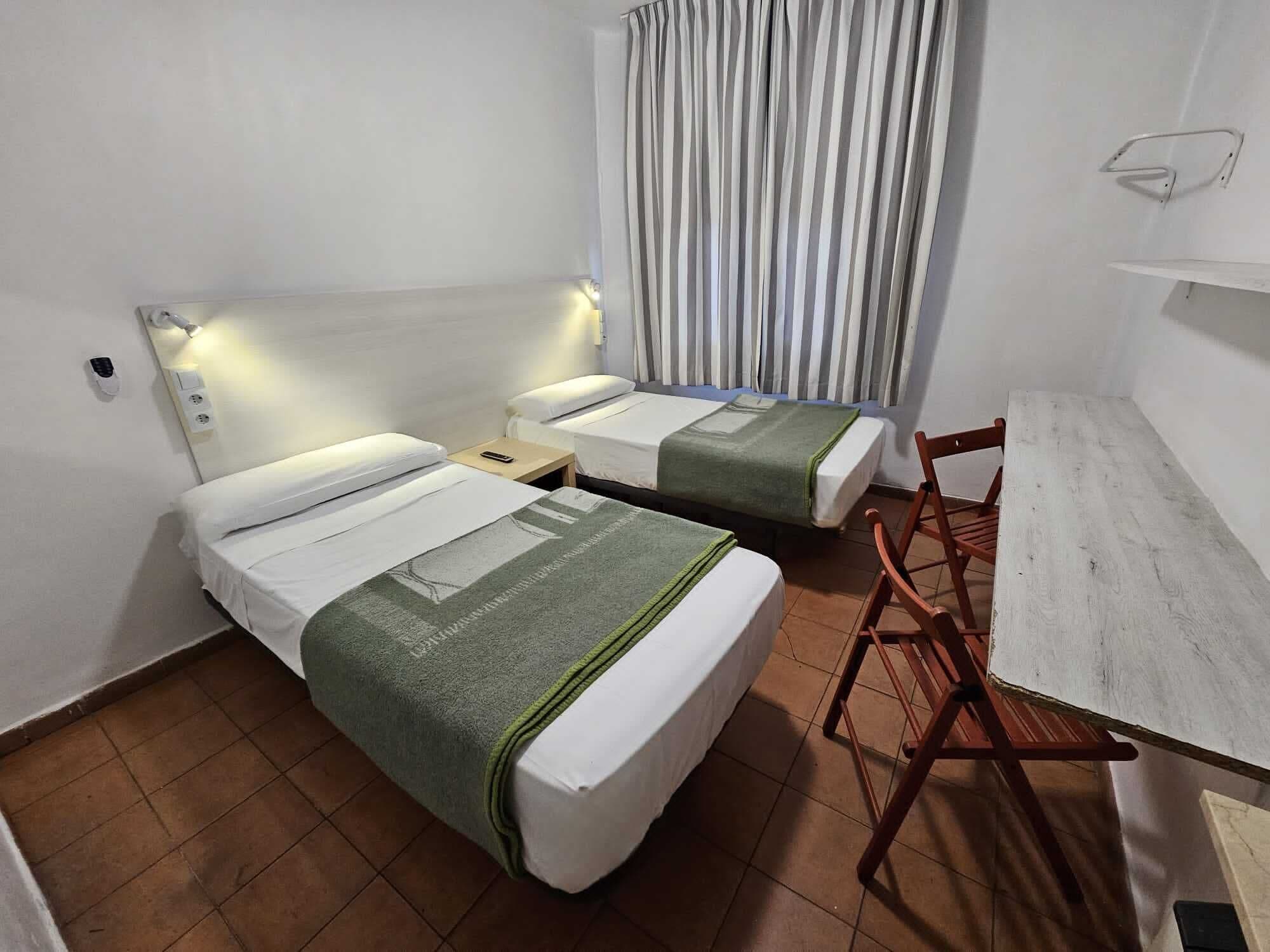 A hotel room with two single beds, a small table, a chair, and a curtain.