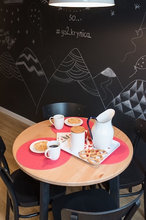Una mesa redonda de madera con un posavasos rojo, una jarra blanca, una taza blanca, una taza de café blanca y un plato con palitos de pretzel.