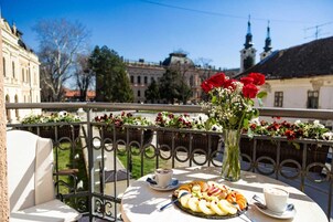 Uma varanda com uma mesa posta para refeição, um vaso com flores e vista para edifícios históricos.