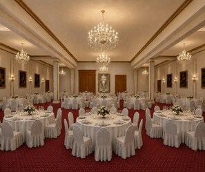 Un salón de banquetes con mesas redondas, sillas blancas, candelabros y cuadros en las paredes.
