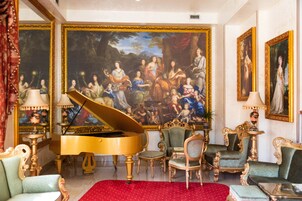 Uma sala imponente com um piano de cauda, cadeiras ricamente decoradas e um grande quadro clássico na parede.