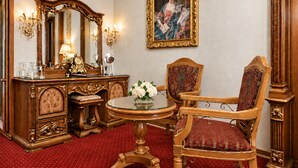 Ein Zimmer mit einem Holzkapuzen-Kleiderschrank, einem Spiegel, einem runden Tisch mit Blumen in einer Vase, einem rot gepolsterten Sessel und einem gerahmten Gemälde an der Wand.