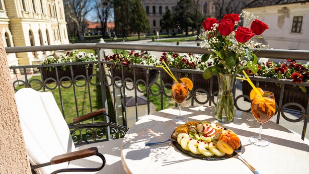 A balcony with a table set for a meal, a vase with red roses, and a view of historic buildings. - Premier Prezident Garni Hotel and Spa (Sremski Karlovci)
