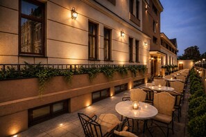 An outdoor seating area with tables and chairs, a building facade with windows, and a warm ambient light.