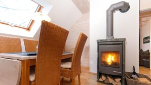 Ein gemütliches Zimmer mit einem Holztisch, Korbstühlen, einem Kamin mit Holzscheiten und einem Fenster mit Aussicht.