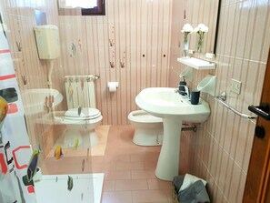Ein Badezimmer mit Toilette, Bidet und Waschbecken. Ein Handtuchhalter und ein kleines Regal mit Blumen sind vorhanden.