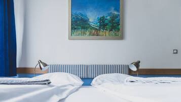 Un lit bien fait, avec du linge de lit blanc, une tête de lit à rayures et deux lampes de chevet. Un tableau encadré représentant une scène de forêt est accroché au-dessus du lit.