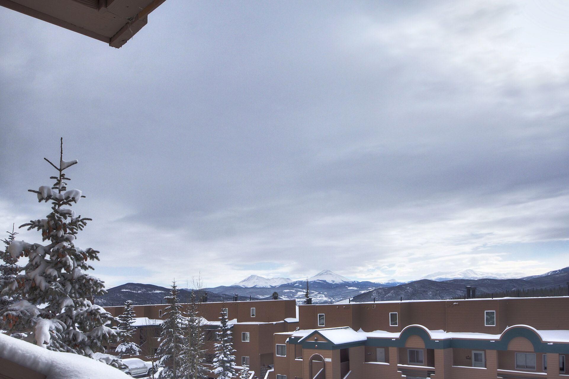 A view of a building complex with multiple brick structures, some with snow on the roofs, and a backdrop of snow-covered mountains under a cloudy sky.