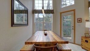 A dining area with a wooden table and benches, large windows with curtains, and a view of trees outside.