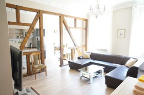 A modern living room with a sectional sofa, a glass coffee table, and a TV. There is a wooden-framed window and a chandelier. - 3 wunderschöne Ferienwohnungen Kornhoff (Naumburg)