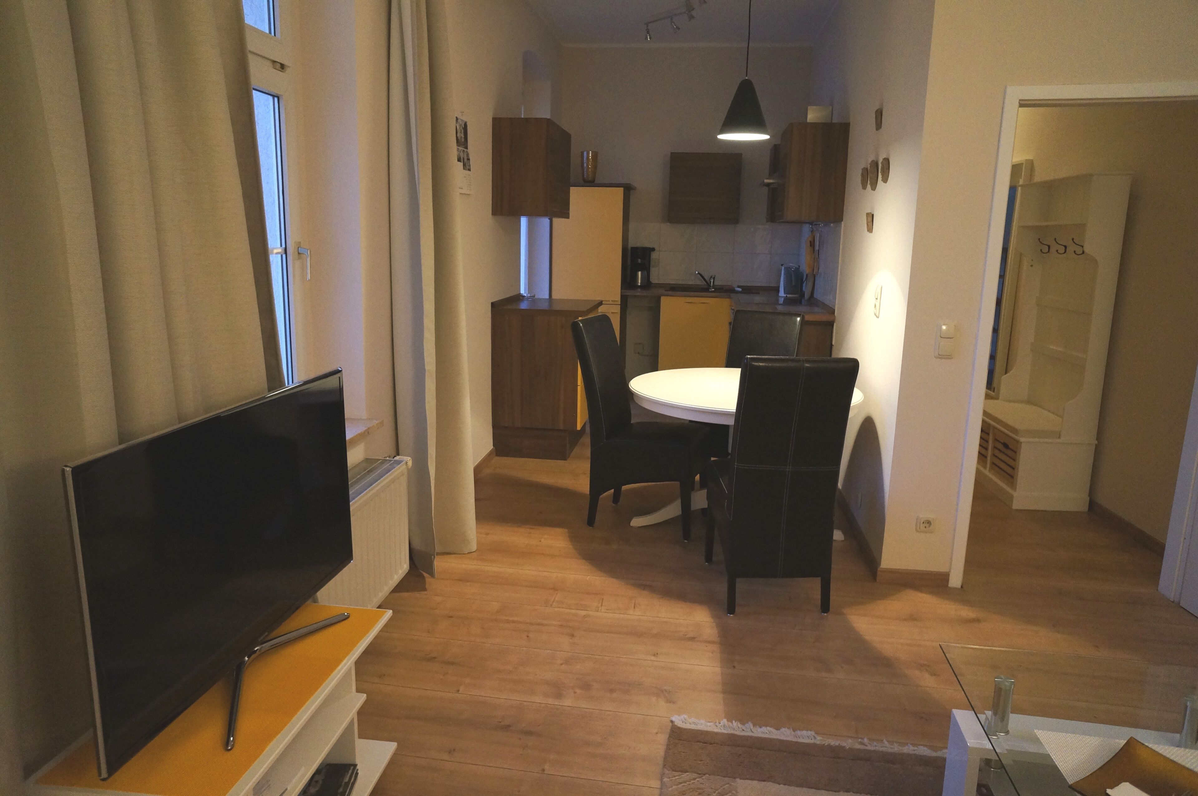 A modern kitchen with wooden cabinets, a dining area with a white table and chairs, and a flat-screen TV on a wooden stand.