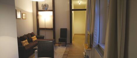 Habitación de hotel con sofá, una mesa de comedor, una silla, televisor y un ventanal grande con cortinas.