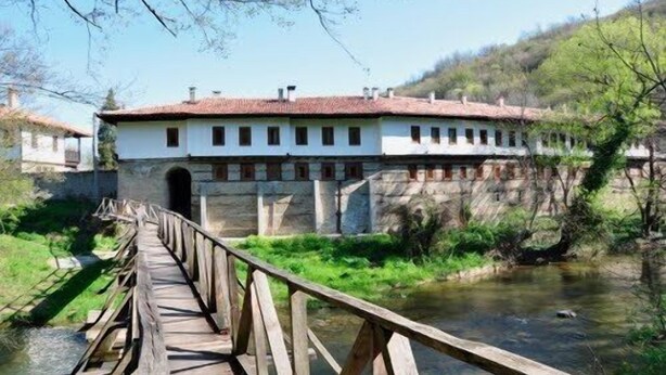 A wooden bridge leading to a white building with a red roof, situated beside a river. - Villa Kalin (Veliko Tarnovo)