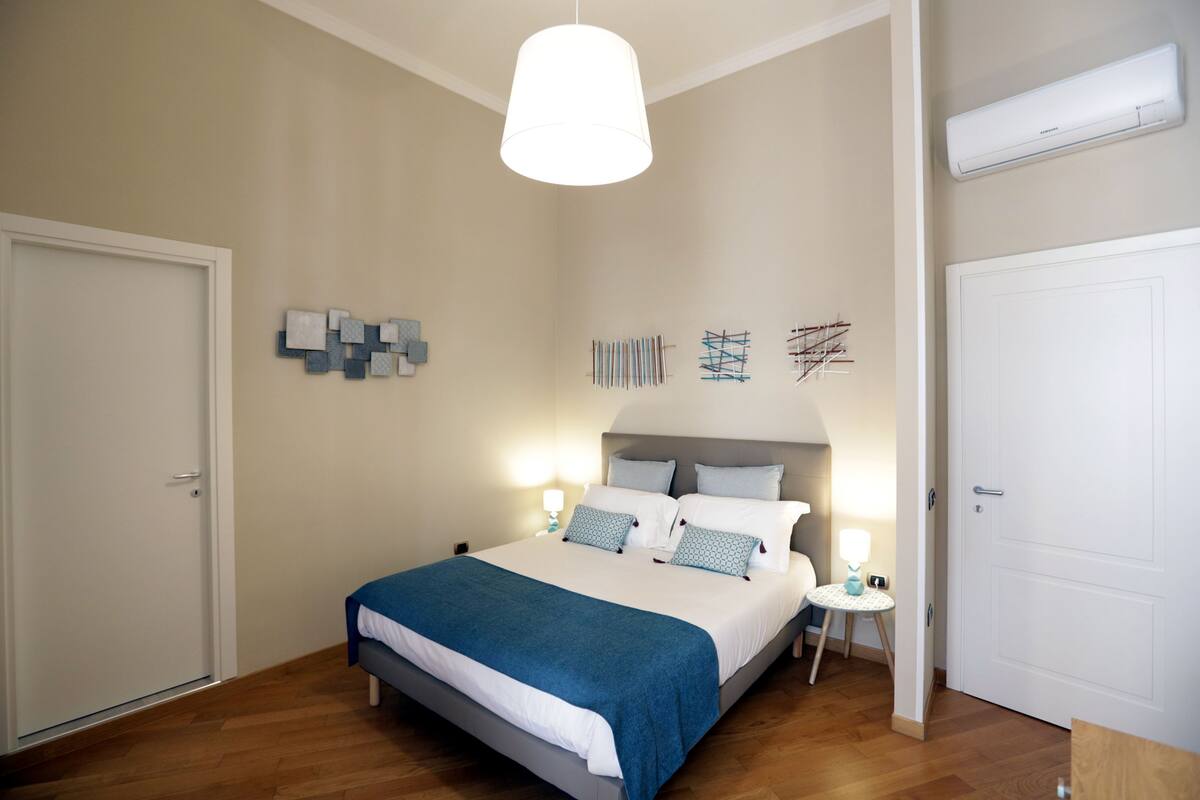 A bedroom with a bed, bedside tables, a hanging light fixture, and a wall-mounted air conditioner.