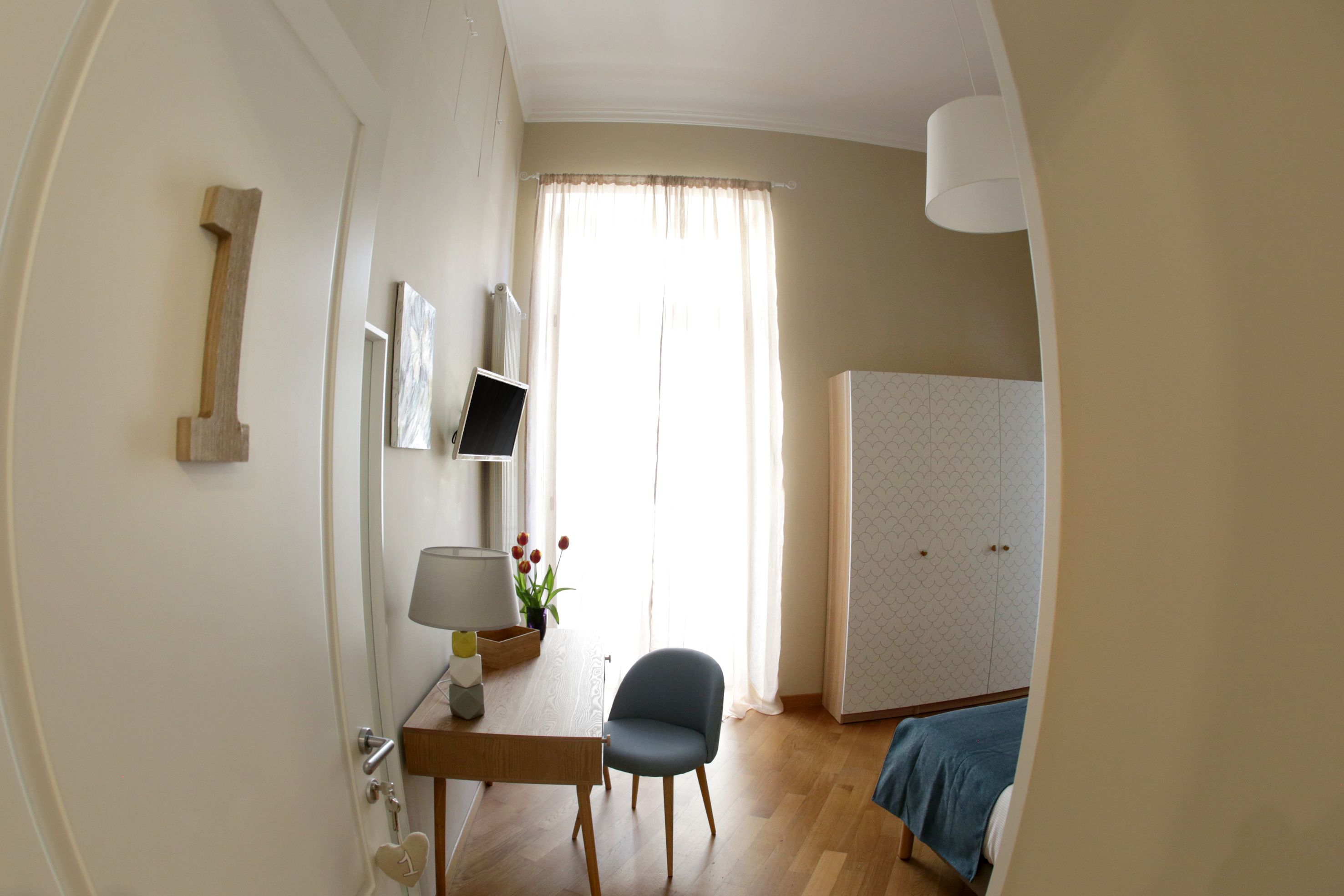 A room with a wooden desk, a chair, a bed with a blue blanket, a wardrobe, and a window with curtains.
