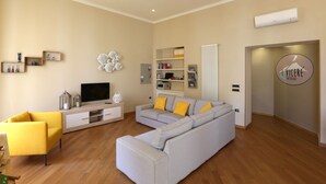 A modern living room with a grey sectional sofa, a yellow armchair, and a flat-screen TV. There are shelves with books and decorative items, and an air conditioning unit on the wall. - I Viceré B&B Deluxe (Naples)