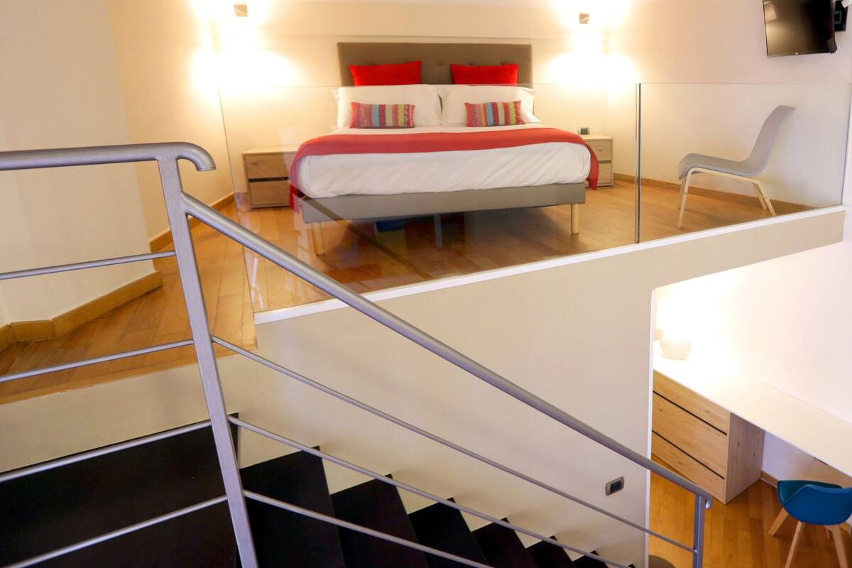 A bedroom with a bed, bedside tables, a desk, and a chair. There is a staircase leading to another level.