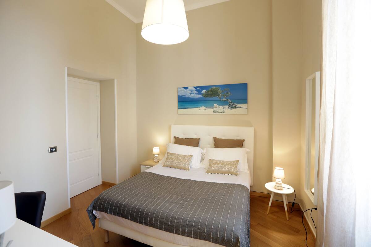 A bedroom with a bed, bedside lamps, a painting of a beach scene, and a door.