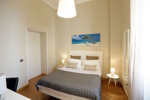 A bedroom with a bed, bedside lamps, a painting of a beach scene, and a door.