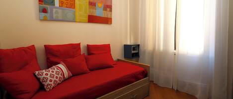 A room with a bed, red bedding, a wall art piece, a nightstand, and a window with curtains.