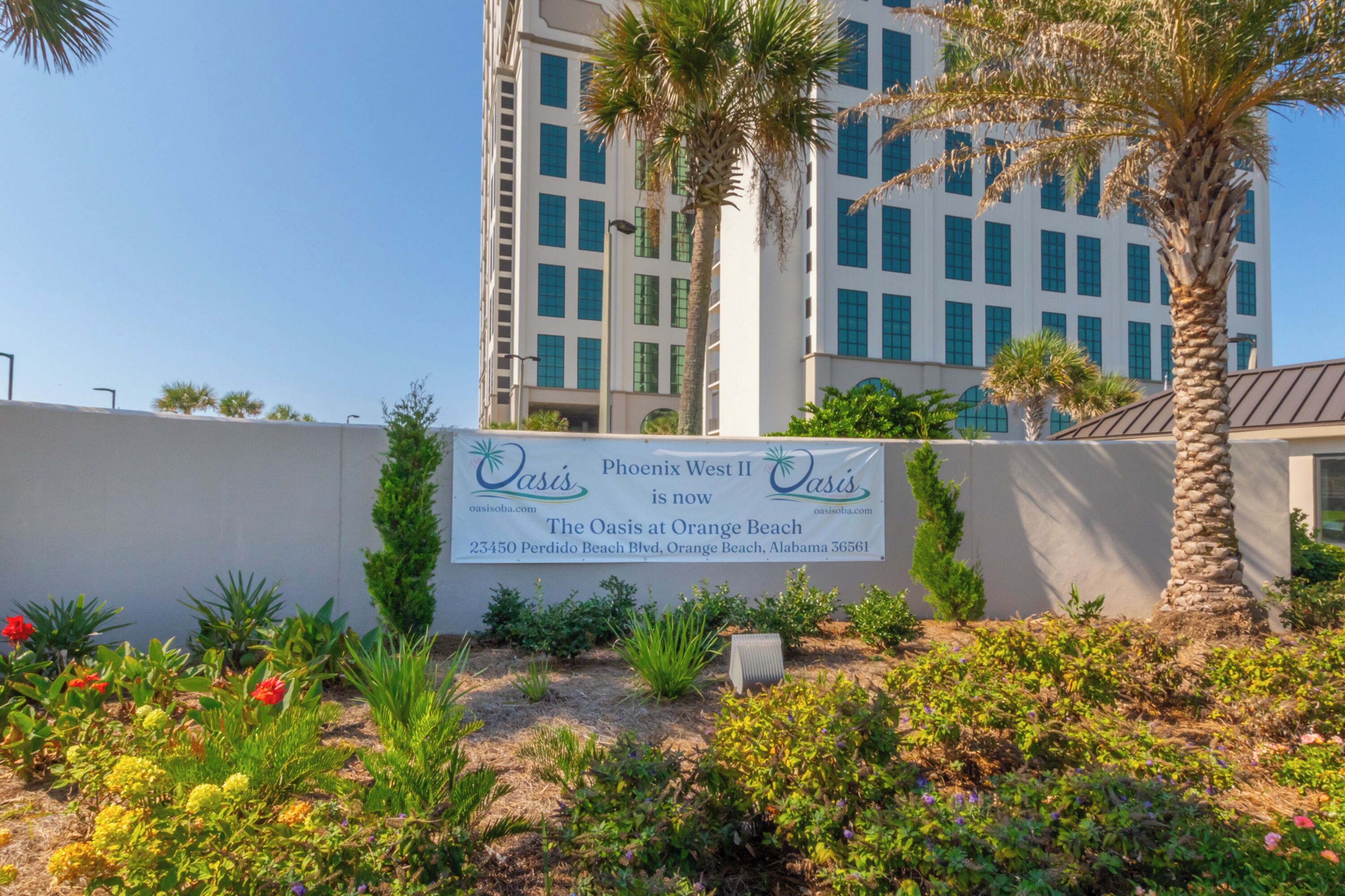 A sign for Oasis at Orange Beach, Phoenix West II, with a view of a tall building and palm trees in the background.
