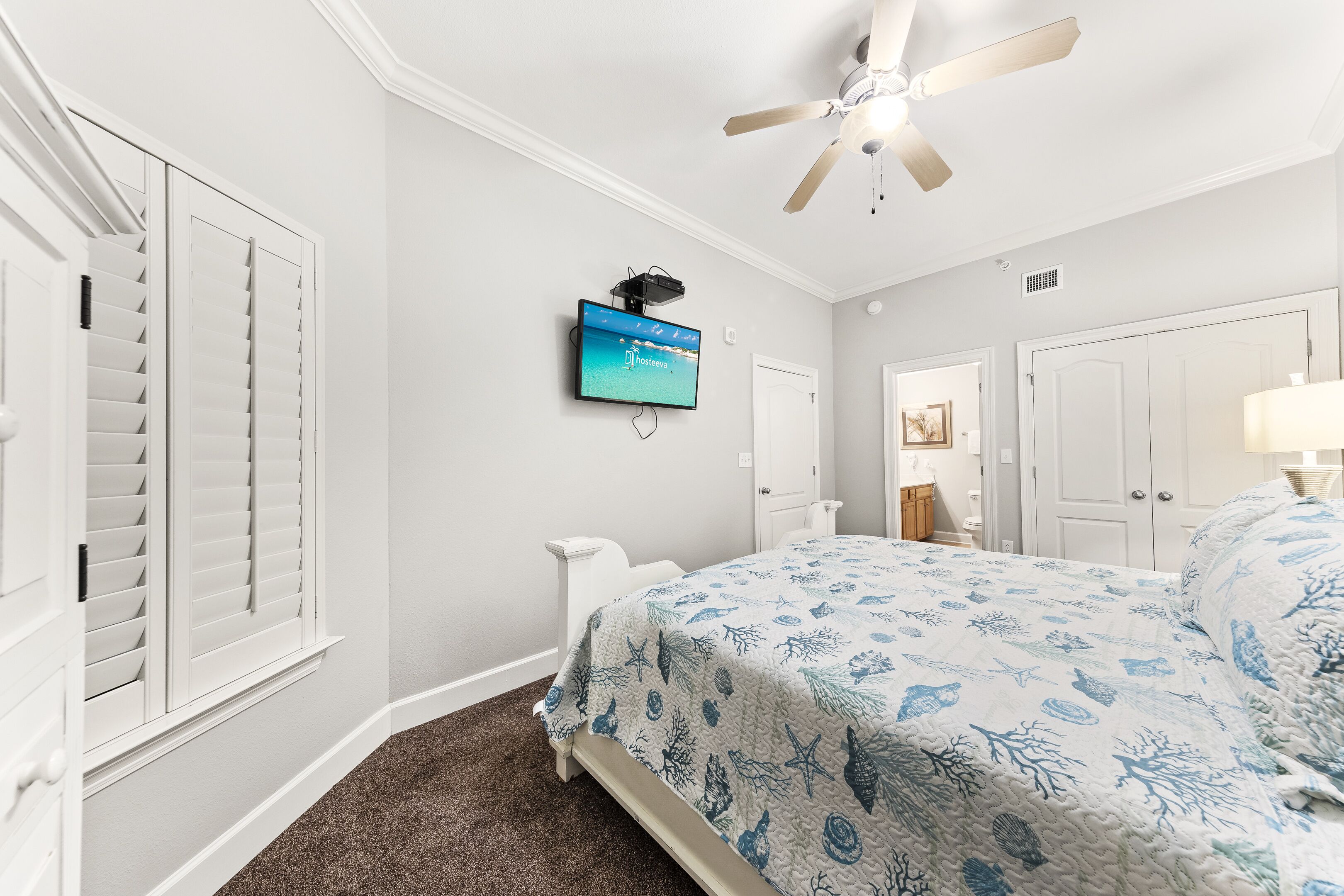 Un dormitorio con cama, ventilador de techo, televisor montado y una puerta con contraventanas blancas.