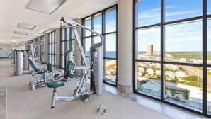 A gym with exercise equipment and large windows overlooking a cityscape. - The Oasis at Orange Beach by Hosteeva  (Orange Beach)